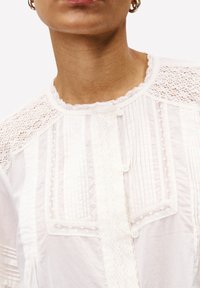 Frau trägt eine weiße Spitzenbluse mit Knopfdetails und goldenen Hängeohrringen, gezeigt vom Hals bis zur oberen Brust.