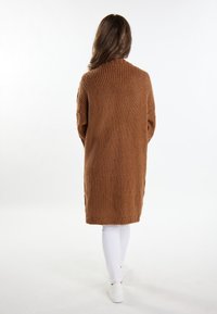 Lang, strikket cardigan i en rig brun farve, der har et tekstureret mønster med en løs pasform og spalte detaljer i siderne. Kombiner med hvide bukser.