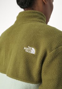 Fleecejacka i olivgrön med en ljusgrön nedre del. Har en hög krage och vit broderad logotyp på vänster bak.