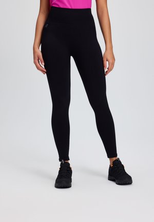 Frau trägt schwarze, hoch taillierte Leggings und schwarze Wanderschuhe, steht vor einem schlichten hellen Hintergrund.