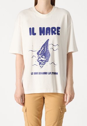 Vrouw die een wit T-shirt draagt met een blauwe schelpafbeelding en Italiaanse tekst "IL MARE" en "LE ONDE INDICANO LA STRADA" met beige cargobroek.