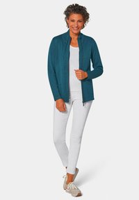 Türkisfarbene Reißverschluss-Strickjacke mit hohem Kragen, gerippter Textur und zwei vorderen Taschen, kombiniert mit weißen Hosen und beigen Sneakers.