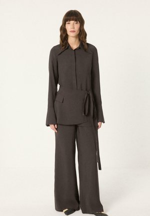 Kobieta stojąca, ubrana w ciemnobrązową bluzkę z długim rękawem typu wrap z szerokimi klapami i dopasowane szerokie spodnie na jednolitym jasnym tle.