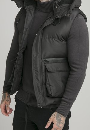 Hombre con chaleco acolchado negro sobre un suéter de cuello alto gris oscuro, con manos tatuadas visibles contra un fondo neutro.