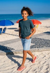 Fille aux cheveux bouclés, portant un t-shirt vert foncé et un short en jean, marchant pieds nus sur une plage de sable près de parasols colorés et de l'océan.