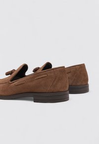 Mocasines de ante marrón con un diseño elegante, que presentan acentos troquelados y una suela plana de goma. Enfocados en la textura y la simplicidad.