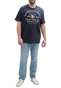 T-shirt à manches courtes bleu marine avec un texte graphique blanc et doré "TOM TAILOR" et "1962" sur la poitrine, associé à un jean bleu clair et des baskets blanches.