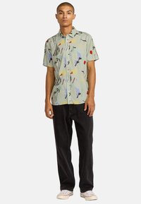 Korte mouwen, lichtgroen knopenoverhemd met een kleurrijk abstract patroon; gecombineerd met zwarte broek en witte sneakers.