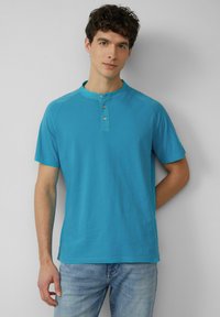 Turkusowa koszulka henley z krótkim rękawem, okrągłym kołnierzykiem i trzema guzikami, wykonana z miękkiego materiału, w połączeniu z jasnoniebieskimi dżinsami.