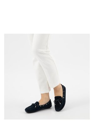 Persoon draagt witte broek en zwarte mocassin loafers met strikdetail, staand op een witte achtergrond.