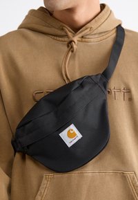 Marsupio in tessuto nero con forma curva, chiusura zip e una prominente toppa con logo arancione e bianco. Indossato sopra un hoodie beige.