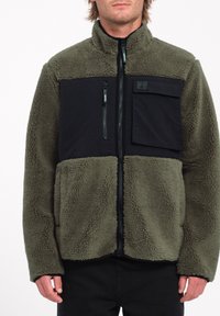 Chaqueta de forro polar con cuerpo verde olivo, panel del pecho negro, bolsillo con cremallera y tejido texturizado. Cuello alto y cierre de cremallera completo.