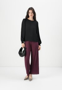 Svart langermet blouse med elastiske mansjetter, paired med plommefargede, plissert vide bukser. Modellen holder en svart håndveske; beige leiligheter fullfører antrekket.