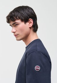 Hombre joven con cabello oscuro, vistiendo una sudadera azul marino con un parche de logo redondo "Colmar" en la parte superior de la manga, mirando hacia la izquierda sobre un fondo liso.