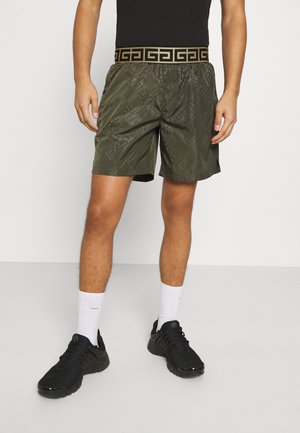 Man met zwarte sneakers, witte sokken, donkergroene korte broek met een zwart-bruine patroon tailleband en een zwart topje.