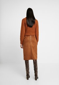 Hnědá semišová midi sukně se zadním zipem, doplněná o texturovaný oranžový pletený svetr, nošená s vysokými botami se vzorem hadí kůže.