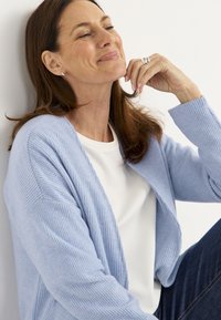 Vaaleansininen ribbipintaista neuletta, joka on valmistettu pehmeästä materiaalista, ja se on puettu valkoisen topin päälle. Omistaa rennon istuvuuden avonaisella edessä ja pitkät hihat.