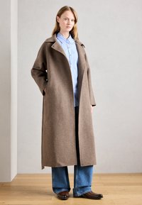Femme portant un long manteau en laine brun sur une chemise bleue claire à boutons, un jean bleu à jambes larges, et des chaussures en cuir brun, se tenant à l'intérieur.