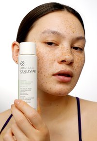 Lotion z kwasem salicylowym Collistar w białej cylindrycznej butelce z zielonymi akcentami. Modelka z piegowatą skórą trzyma butelkę obok twarzy.