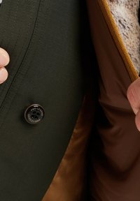 Groene gestructureerde stof met een verticale streepjespatroon, een ronde zwarte knop gemarkeerd "PME-LEGEND," en een zichtbaar bruin satijnen voering.