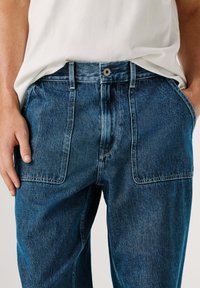 Tumesinised teksapüksid klassikalise disainiga, nähtavate õmblustega, kahe esitasku ja vöökohal nööpidega sulgemine.