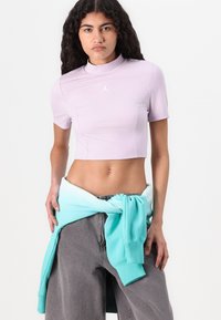 Világoslila rövid ujjú, magas nyakú cropped top, apró fehér logóval. Szürke nadrággal és derékon megkötött türkizkék pulóverrel párosítva.