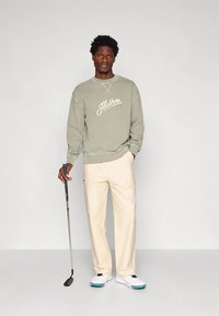 Lichtgroene trui met witte logotekst, gecombineerd met crèmekleurige broek en witte golfschoenen met blauwe accenten, terwijl hij een golfclub vasthoudt.