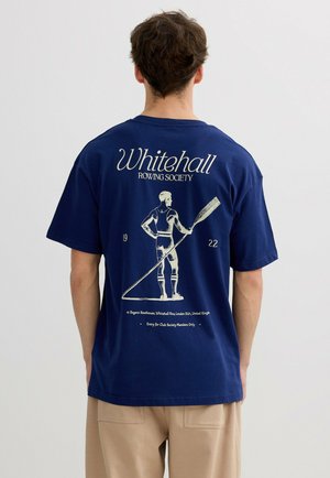 Hombre con camiseta azul marino con gráfico y texto de estilo vintage de Whitehall Rowing Society en la parte posterior, combinado con pantalones beige.
