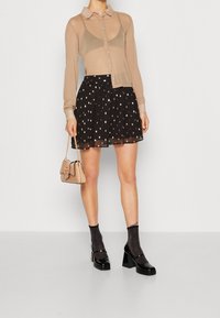 Chemise beige transparente à boutons sur une jupe plissée noire à pois dorés, accompagnée de chaussures à talons noires et de chaussettes scintillantes, tenant un petit sac à main.
