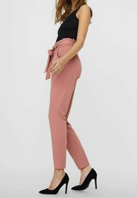 Pantalon rose sur mesure avec ceinture, taille haute et jambes fuselées. Porté avec un haut noir et des chaussures à talons hauts noires. Texture lisse.