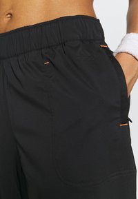 Zbliżenie na osobę w czarnych szortach Adidas z elastycznym pasem, trzymającą rękę w kieszeni, z nadgarstkiem owiniętym białą opaską.