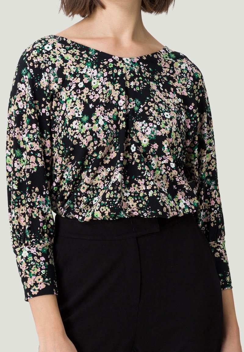 Femme portant un chemisier noir à manches longues avec un petit motif floral rose, blanc et vert, rentré dans un pantalon noir taille haute.