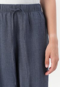 Pantalon gris texturé avec une taille élastique et un cordon de serrage. Le tissu lisse présente un motif en grille subtil. Main reposant à côté du tissu.