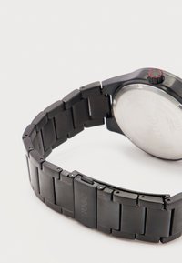 Μαύρο μεταλλικό λουράκι ρολογιού με αγκράφα χαραγμένη με τη λέξη "HUGO" και μερικώς ορατή στρογγυλή θήκη ρολογιού με κόκκινο δακτύλιο γύρω από το κουμπί, σε λευκό φόντο.