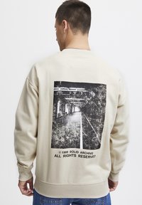 Sudadera beige con un gran gráfico en blanco y negro desgastado de un túnel del metro en la parte posterior. El texto en la parte inferior indica detalles de copyright.