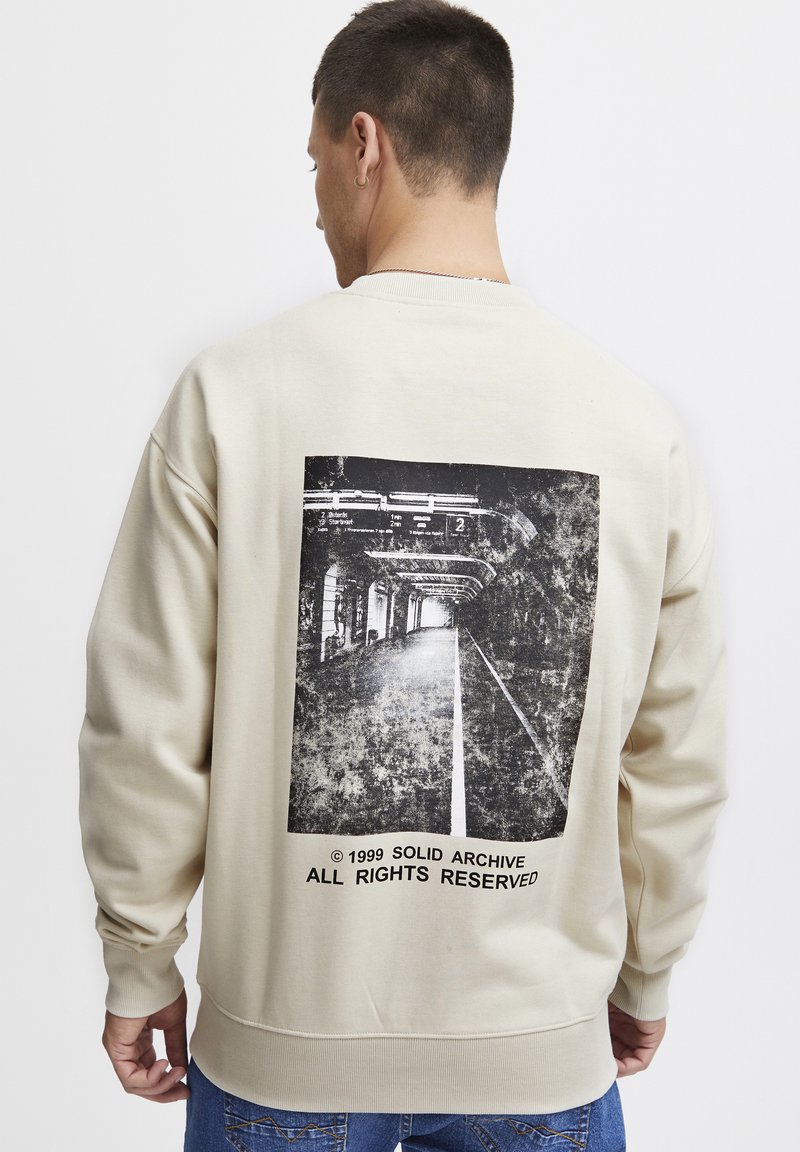 Sudadera beige con un gran gráfico en blanco y negro desgastado de un túnel del metro en la parte posterior. El texto en la parte inferior indica detalles de copyright.