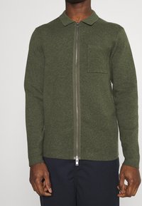 Man som bär en olivgrön zip-up cardigan med krage och bröstficka, tillsammans med mörka byxor.