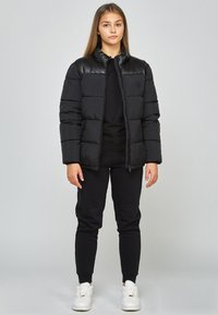 Svart puffjacka med en glänsande övre del, dragkedjestängning och sidofickor, i kombination med svarta joggers och vita sneakers.