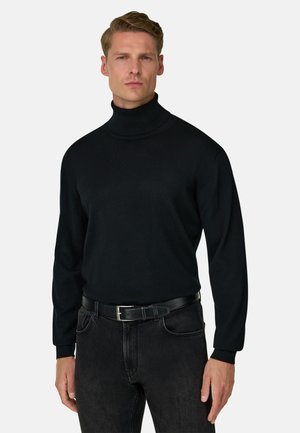 Homme portant un pull à col roulé noir rentré dans un jean noir avec une ceinture noire, debout devant un fond clair uni.