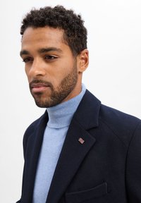 Marineblauer Blazer kombiniert mit einem hellblauen Rollkragenpullover. Der Blazer hat eine Brusttasche und eine kleine Union-Jack-Nadel am Revers.