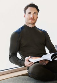 Olivenfarvet langærmet bluse med marineblå detaljer, tætsiddende design og høj krave. Model siddende med en bog mod en hvid baggrund.