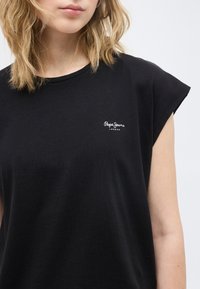 Γυναίκα με ξανθά μαλλιά μήκους μέχρι τους ώμους, φορώντας μαύρο αμάνικο τοπ με το λογότυπο "Pepe Jeans London" στο στήθος.