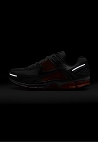 Black athletic shoe with a mesh upper, reflective accents, and orange sole. Features a textured design with a curved shape and padded collar.