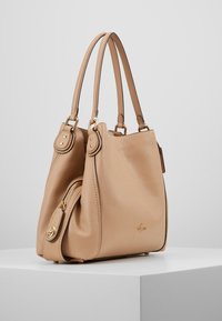 Sac à main en cuir beige avec une texture lisse, des poignées doubles et des accessoires en métal doré. Il dispose d'une poche latérale zippée pour un espace de rangement supplémentaire.