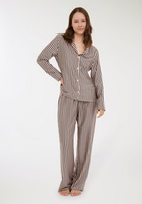 Donna in piedi a piedi nudi, con una camicia a maniche lunghe beige e nera a righe verticali e pantaloni larghi coordinati.