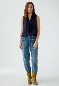Navyfarbener ärmelloser Top mit Schleifendetail, kombiniert mit hellblauen hochgeschnittenen Jeans. Das Model trägt beigefarbene Wildleder-Ankle-Boots.