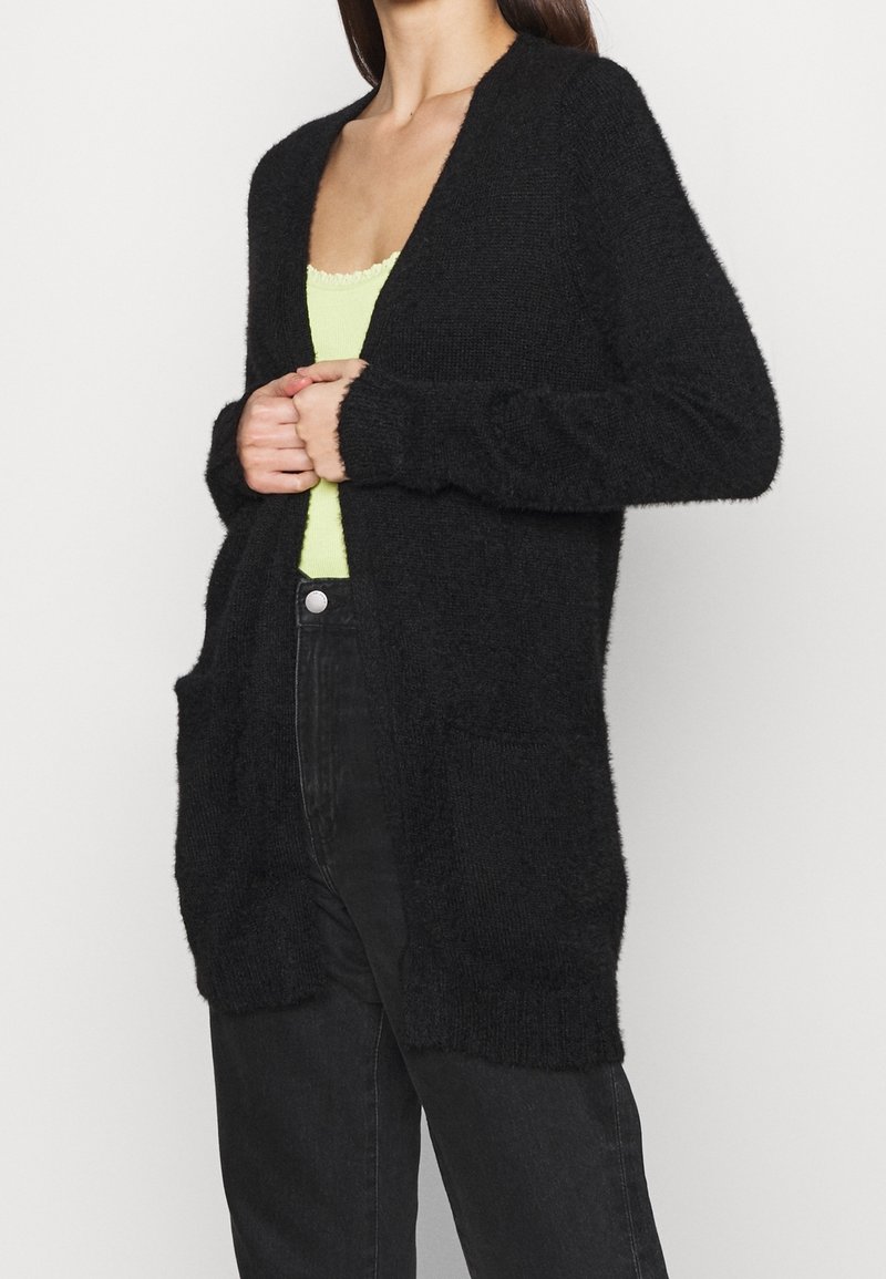 Kvinna iklädd en svart fluffig cardigan, en neongrön linne och högmidjade svarta jeans, händerna ihopvikta vid cardiganens öppning.