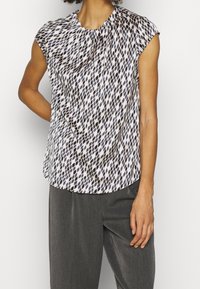 Bluzka z krótkim rękawem w białym kolorze z geometrycznym czarno-beżowym wzorem, z zaokrąglonym dekoltem i miękką, lejącą się tkaniną.
