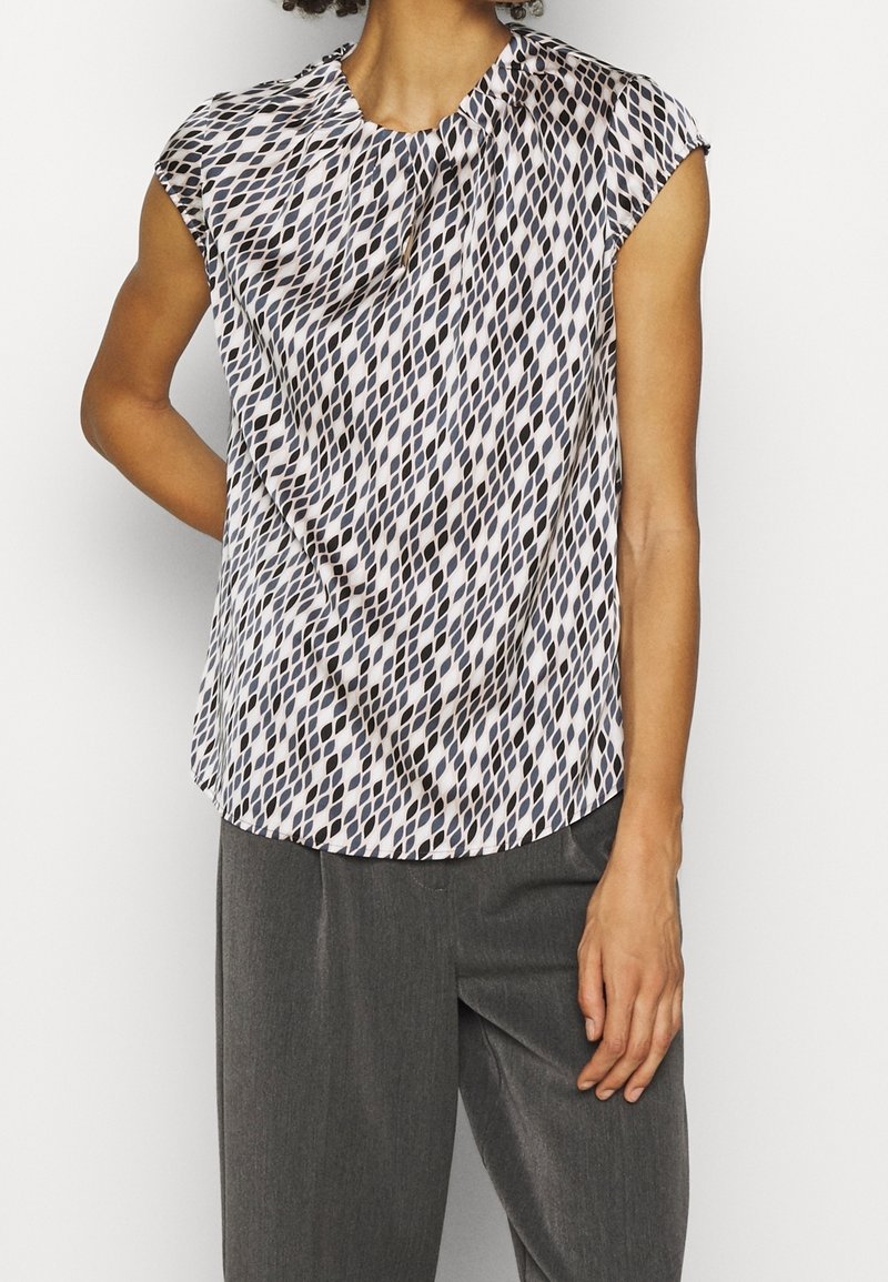 Bluzka z krótkim rękawem w białym kolorze z geometrycznym czarno-beżowym wzorem, z zaokrąglonym dekoltem i miękką, lejącą się tkaniną.