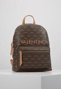 Mochila marrón de lona recubierta con patrón de logo en relieve. Presenta un cierre de cremallera dorada, un bolsillo frontal y detalles en cuero beige.